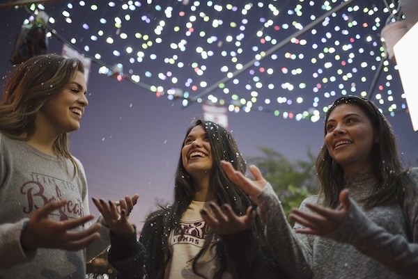 Students wearing Texas State shirts surrounded by holiday lights and catching artificial snow