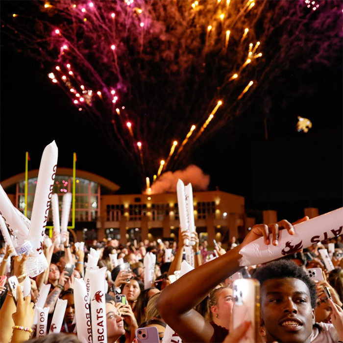 Students on the field at UFCU stadium with fireworks in the sky